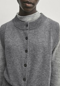 Close-up of a person wearing a gray buttoned cardigan over a light gray turtleneck sweater against a plain background.