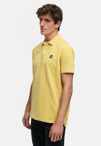 Gul pikétröja i bomull med krage, tre knappar och en avvikande svart rund logotyp på bröstet.