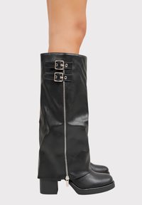 Bottes noires montant jusqu'au genou avec une finition en cuir brillant, dotées de deux boucles argentées et d'une fermeture éclair sur le côté, avec un bout carré et un talon bloc.