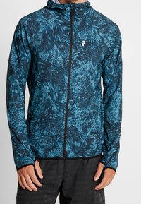 Veste légère à capuche avec un motif abstrait bleu et noir, dotée d'une fermeture éclair intégrale et d'ouvertures pour les pouces aux poignets pour un maintien sécuritaire.