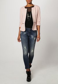 Vaaleanpunainen bleiseri, jossa on pyöreä kaula-aukko, musta verkkopaita alla, kuluneet siniset farkut ja mustat nilkkurit, joissa on hihnat.