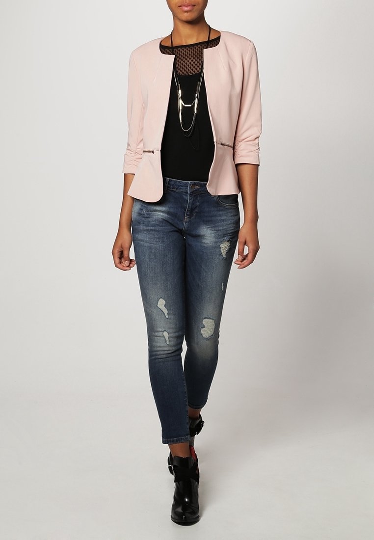 Vaaleanpunainen bleiseri, jossa on pyöreä kaula-aukko, musta verkkopaita alla, kuluneet siniset farkut ja mustat nilkkurit, joissa on hihnat.