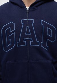 Personne portant un sweat à capuche zippé bleu marine avec de grandes lettres contour "GAP" sur la poitrine et des poches avant sous la fermeture éclair.