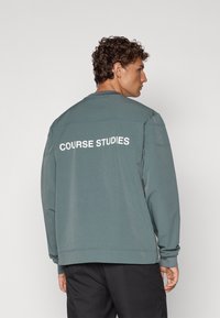 Grå hettegenser med rund hals og lange ermer, med teksten "COURSE STUDIES" i hvitt på ryggen. Myk stoffkvalitet og løs passform.