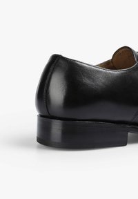 Zapato de cuero negro con acabado pulido, tacón redondeado y suela negra resistente. Presenta una textura suave y un diseño clásico.