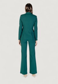 Suit sartorial teal con giacca aderente e pantaloni svasati. Tessuto liscio con cuciture strutturate, silhouette posteriore visibile.