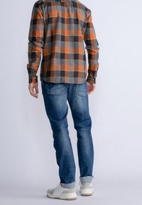 Koszula w kratę w kolorach pomarańczowym, szarym i czarnym; zestawiona z niebieskimi jeansami i białymi tenisówkami. Widok od tyłu pokazuje długość i dopasowanie koszuli.