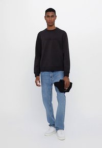 Άντρας που στέκεται, φοράει μαύρη μπλούζα, ανοιχτόχρωμο μπλε τζην, λευκά αθλητικά παπούτσια, κρατώντας μια μικρή μαύρη τσάντα στο δεξί χέρι μπροστά σε απλό φόντο.