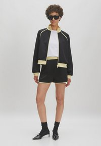 Veste de sport noire avec des accents jaunes, associée à un short assorti. T-shirt blanc en dessous. Mannequin portant des lunettes de soleil et des escarpins.