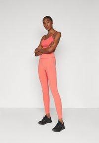 Korall színű sportmelltartó és hozzá illő, magas derekú leggings texturált anyagból, fekete sportcipővel párosítva. Minimális márkázás látható.