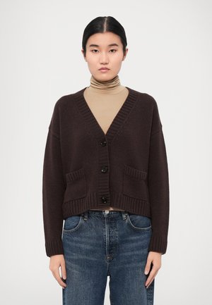Cardigan court marron foncé en tricot texturé, avec un col en V, cinq boutons noirs et deux poches avant. Porté sur un léger col roulé.