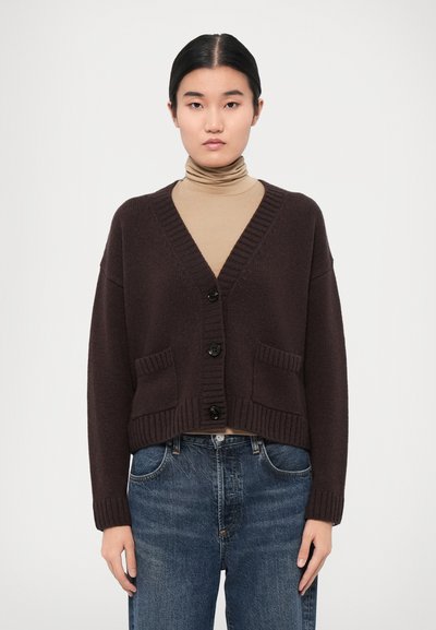 Dunkelbrauner, taillierter Cardigan aus strukturiertem Strick, mit V-Ausschnitt, fünf schwarzen Knöpfen und zwei vorderen Taschen. Trägt man über einem hellen Rollkragenpullover.