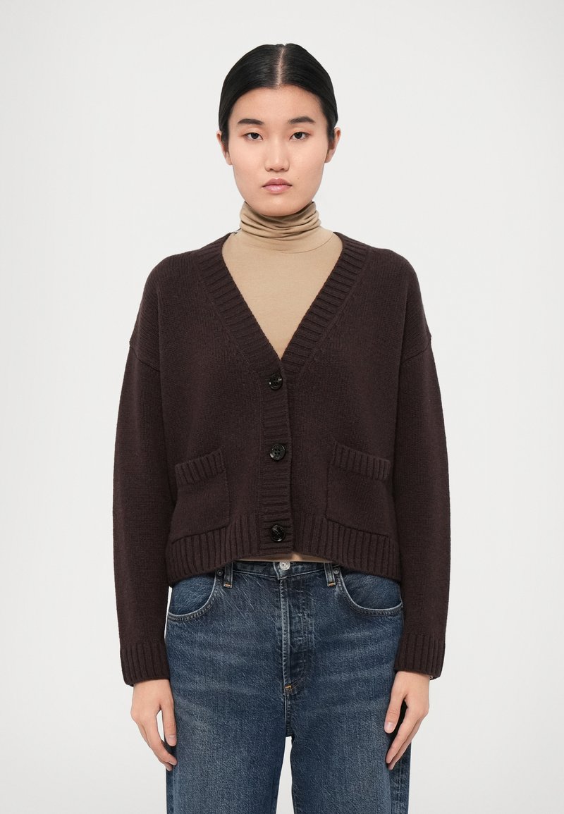 Dark brown cropped cardigan made of textured knit, featuring a v-neck, five black buttons, and two front pockets. Worn over a light turtleneck.