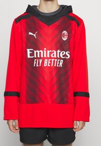 Rotes Fußballtrikot mit langen Ärmeln und Kapuze, mit schwarzen Akzenten und einem Zickzackmuster. Logo auf der Brust und der Text "Emirates Fly Better" prominent angezeigt.