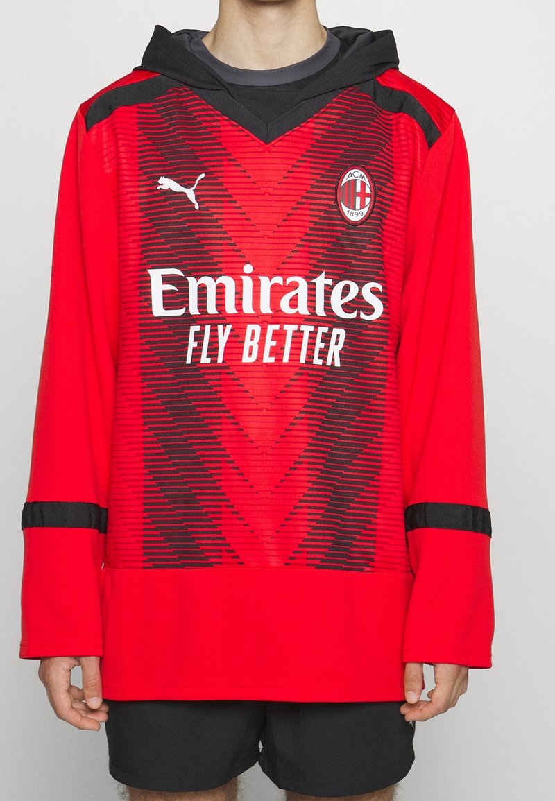 Rotes Fußballtrikot mit langen Ärmeln und Kapuze, mit schwarzen Akzenten und einem Zickzackmuster. Logo auf der Brust und der Text "Emirates Fly Better" prominent angezeigt.