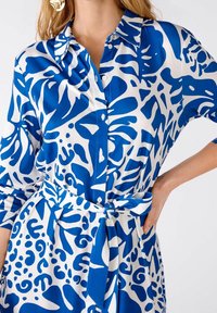 Abito camicia blu e bianco a fantasia, con colletto, chiusura con bottoni, maniche larghe e un accento in vita con nodo. Tessuto dalla texture liscia.