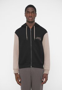 Sudadera con capucha de cremallera negra con mangas beige, cuenta con un bolsillo frontal y capucha con cordón, terminada con un texto bordado en el lado izquierdo del pecho.