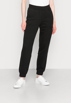 Personne portant un pantalon de jogging noir avec des revers et des baskets blanches, debout devant un fond clair uni.