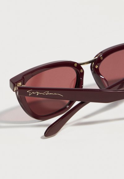 Γυαλιά ηλίου μπορντό σχήμα "cat-eye" με σκουρόχρωμους φακούς και χρυσή υπογραφή στο πλευρό, ακουμπισμένα σε λευκή επιφάνεια.