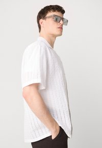 Witte kortebloeiende shirt met gestructureerde stof en verticale strepen, met een knoopsluiting aan de voorkant en een kraag. Gemaakt met grijze zonnebril.