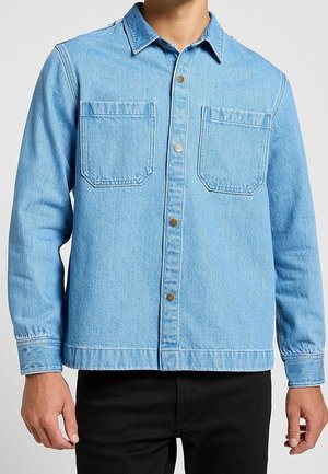 Man draagt een lichtblauw denim blouse met knopen, twee borstzakken en lange mouwen, gecombineerd met een zwarte broek.