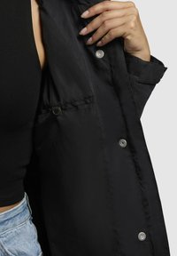 Veste noire avec une texture lisse, dotée de boutons-pression en argent et d'une poche latérale. Col ouvert, présentée avec une main tenant le revers.