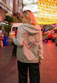 Felpa grigia con una grafica a forma di cuore rosso e design di spilli di sicurezza neri sulla schiena. Il modello tiene una bevanda mentre cammina in una strada vibrante.