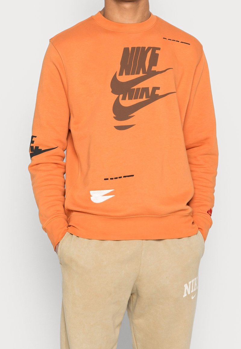 Person som bär en orange Nike-tröja med flera svarta och vita Nike-loggor samt beiga byxor med en vit Nike-logga.