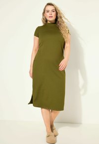 Olivgrünes, geripptes Midikleid mit kurzen Ärmeln, hohem Ausschnitt und seitlicher Schlitz, kombiniert mit beigen Slippern. Glatte Textur.