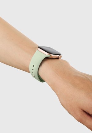 Nadgarstek noszący smartwatch w kolorze różowego złota z czarnym ekranem i jasnookie zieloną silikonową opaską na jednolitym jasnym tle.