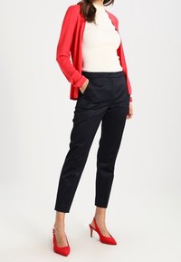 Cardigan rojo sobre una blusa blanca de canalé, combinado con pantalones azul marino con estampado y tacones rojos. El conjunto presenta un diseño ajustado y una forma entallada.