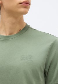 Κοντινό πλάνο ενός ατόμου που φοράει ένα ανοιχτό πράσινο μπλουζάκι EA7 Emporio Armani, με το λογότυπο στο στήθος και μέρος του λαιμού και των χειλιών ορατά.