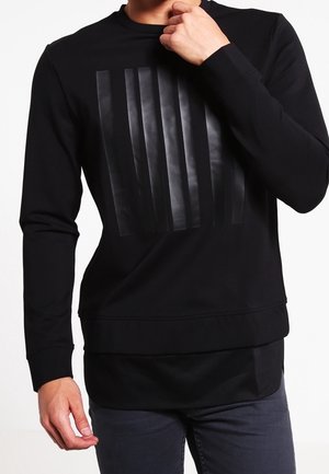 Man draagt een zwart shirt met lange mouwen en glanzende verticale zwarte strepen op de borst, gecombineerd met donkere broek.