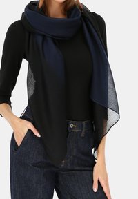 Czarny lekki szalik zarzucony na dopasowany czarny top, zestawiony z ciemnymi dżinsami. Szalik ma płynny wzór i miękką teksturę.