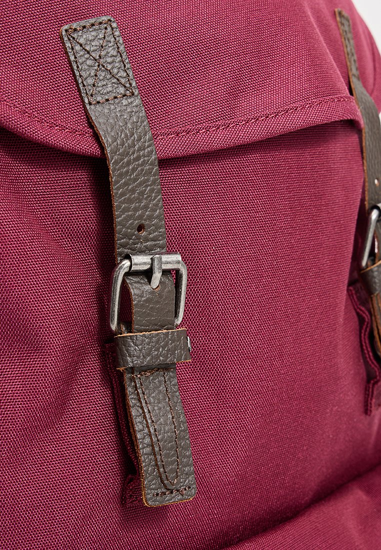 Maronenfarbene Stoff-Rucksack mit braunen Lederträgern und Metall-Schnallen, detaillierter Stickerei und einer strukturierten Oberfläche.