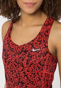 Débardeur de sport rouge et noir à motifs avec un col en U et des bretelles fines, arborant un petit logo blanc sur le devant.