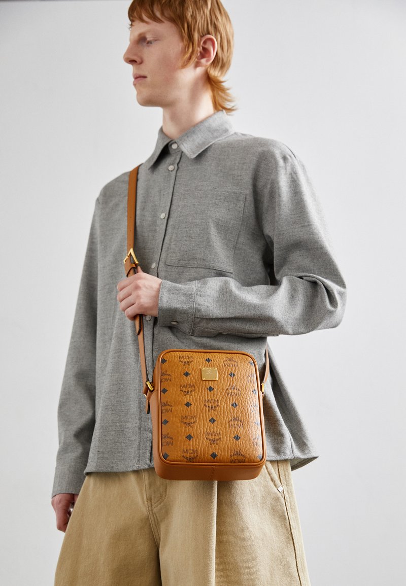 Hnědá kožená kabelka přes tělo s embosovanými vzory, čtvercového tvaru, se zlatými kovovými detaily a odnímatelným ramenním popruhem, nošená s šedou košilí.