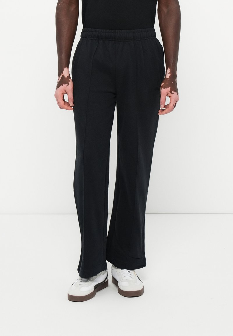 Personne portant un pantalon noir à jambes larges, des baskets blanches avec des semelles marron, debout devant un fond blanc uni.
