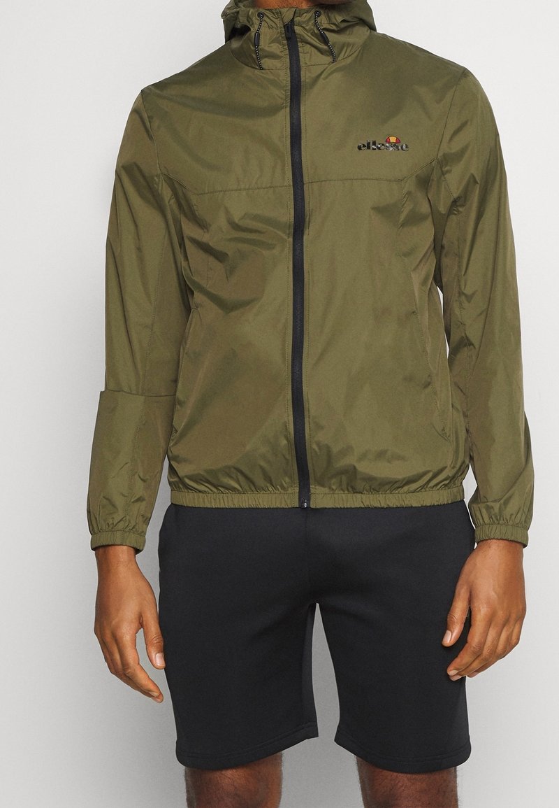 Giacca leggera verde oliva con zip frontale, cappuccio regolabile, polsini elastici e dettaglio con logo sul petto. Abbinata a pantaloni corti neri.
