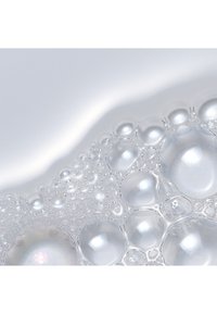 Przezroczysty roztwór piany z bąbelkami o różnej wielkości, odbijającymi światło, na gładkiej, białej powierzchni.