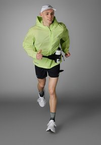 Hellgrüner Windbreaker mit Kapuze, schwarze Shorts und ein Hydrationsgürtel. Weiße Mütze und Schuhe, graue Socken. Läufer in Bewegung vor einem grauen Hintergrund.