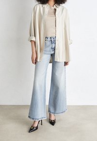 Camicia oversize color beige chiaro, top fitted beige, jeans a gamba larga blu chiaro e scarpe col tacco nere con dettagli dorati.