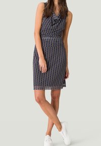 Granatowa sukienka z dekoltem w łódkę, geometryczny wzór w odcieniach kremu i beżu, dopasowana w talii, długość do kolan, zestawiona z białymi sneakersami.
