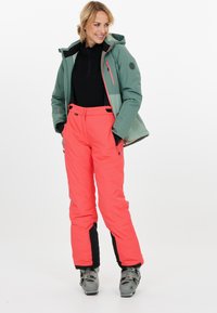 Skijas in teal met een capuchon, voorzien van een ritszak. Felroze skibroek met zwarte accenten en schoenen voor wintersport.