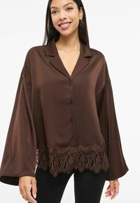 Brun satengbluse med lange ermer, krage, knappelukking og dekorativ blonderkant nederst. Glatt tekstur, løs passform.