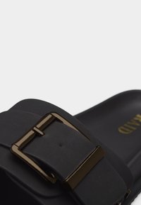 Sandale noire à semelle glissière avec une large bride et une grande boucle métallique rectangulaire brillante, montrant une partie de la semelle intérieure avec des lettres dorées.