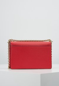 Rektangulær rød lædertaske med en tekstureret overflade, gylden kæderekke og en flad frontplade. Minimalistisk design uden synligt hardware.