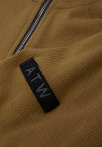 Brauner Fleece-Stoff mit feiner Rippstruktur, ausgestattet mit einem schwarzen Stoffpatch mit der Aufschrift "ATW" und einem silbernen Reißverschluss.