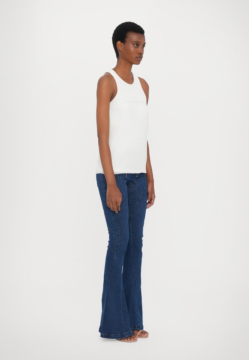 Top sin mangas blanco ligero con escote redondo, combinado con jeans de campana azul oscuro que presentan un diseño de costura diagonal y un hardware mínimo.