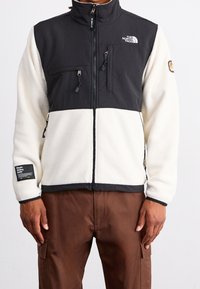 Fleecejacke mit einem schwarzen Oberteil und einem cremefarbenen Unterteil. Verfügt über Reißverschlusstaschen und ein Logopatch am linken Ärmel.